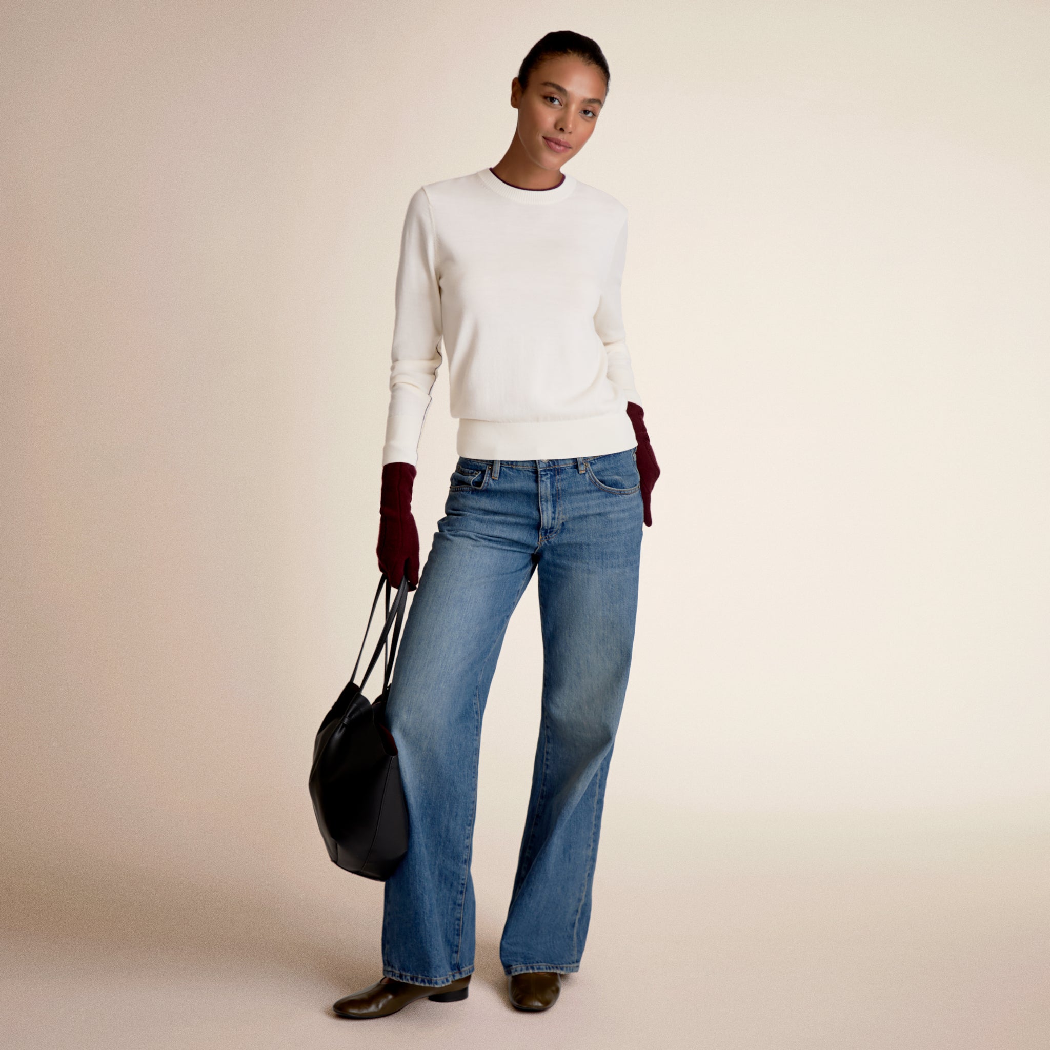 Woman wearing a white sweater and blue jeans, holding a black bag on a beige background