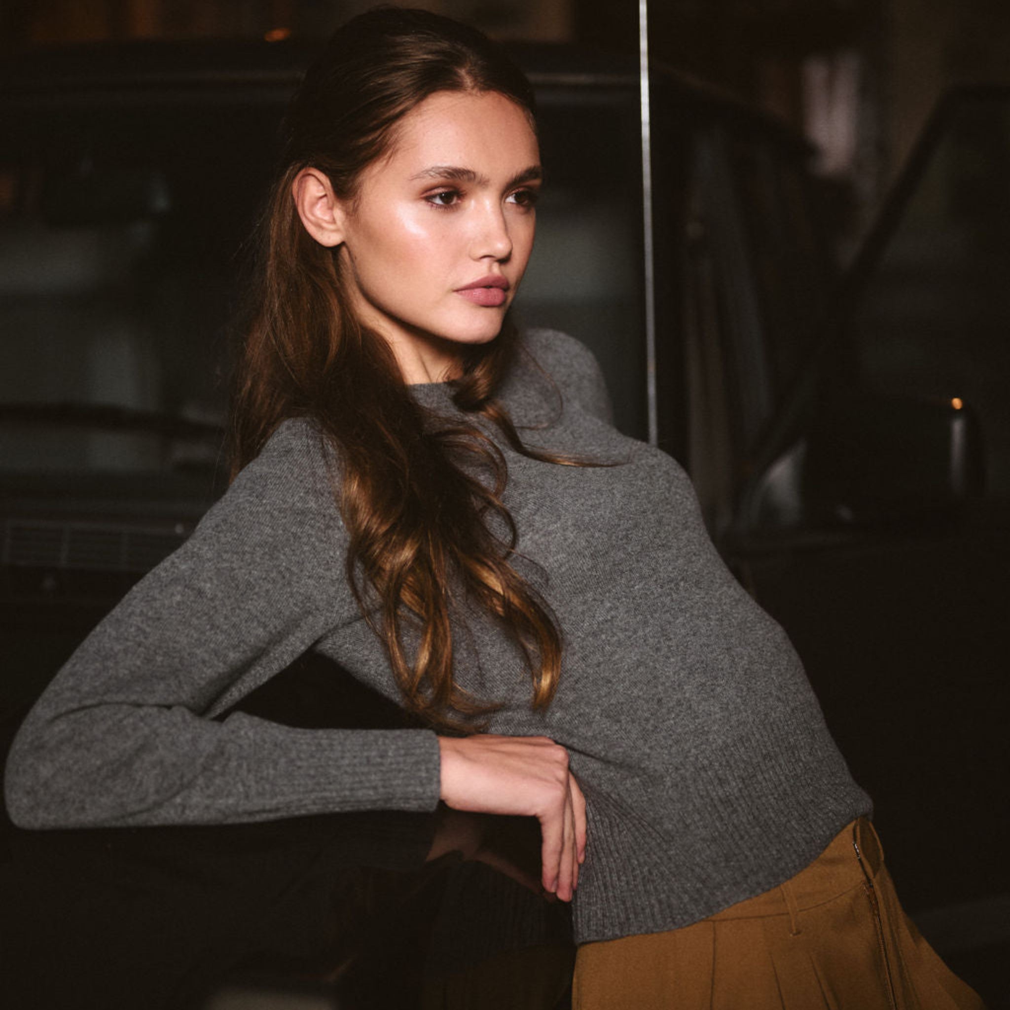 Woman in a gray sweater and brown pants sitting in a car at night. 
