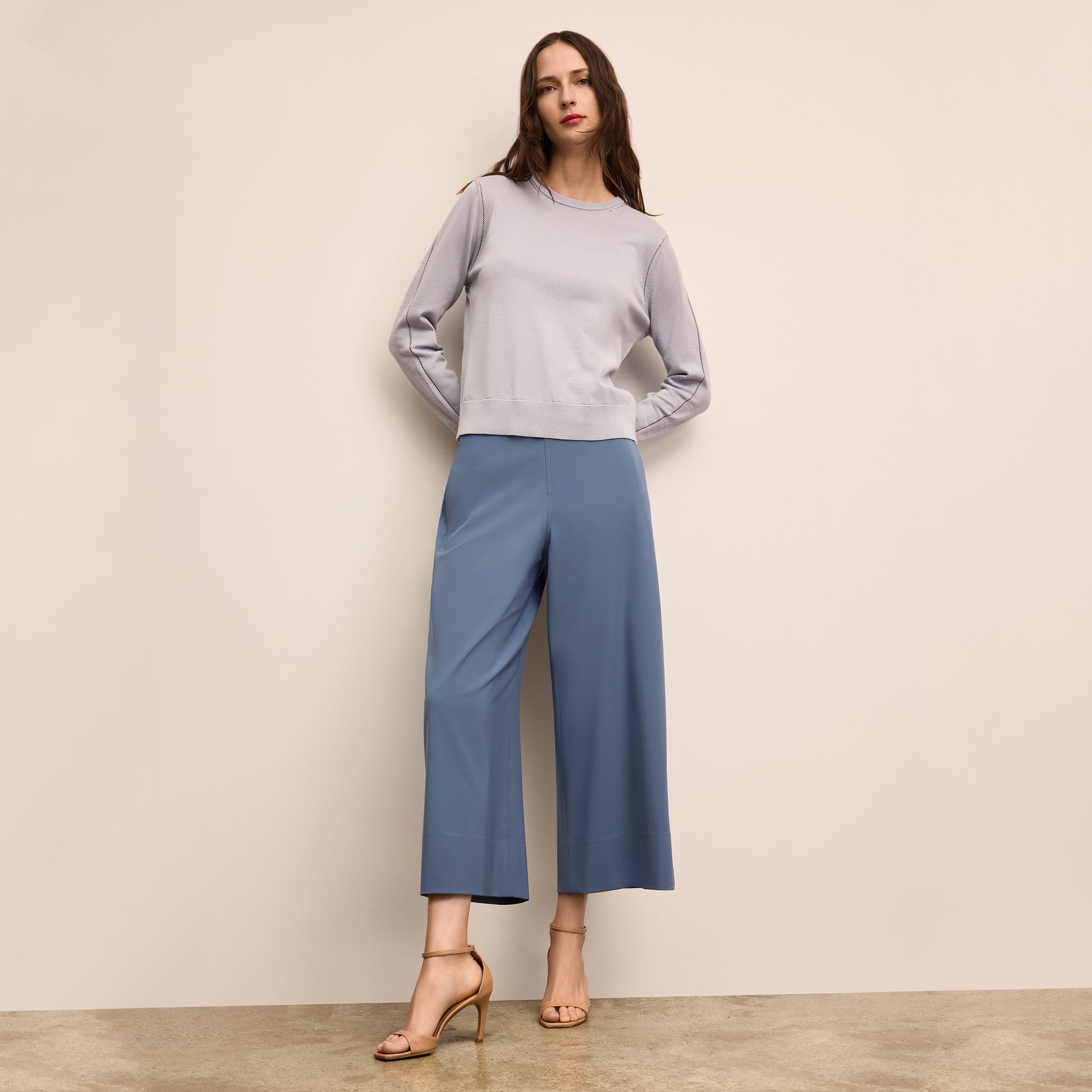 Woman wearing a light gray sweater and blue pants against a beige wall 