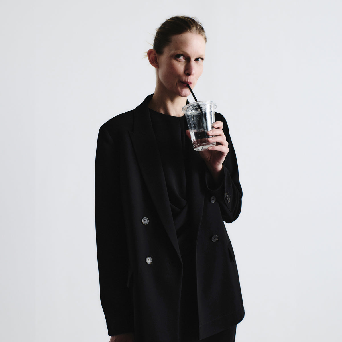 Person wearing a black coat holding a clear cup with a straw against a plain background