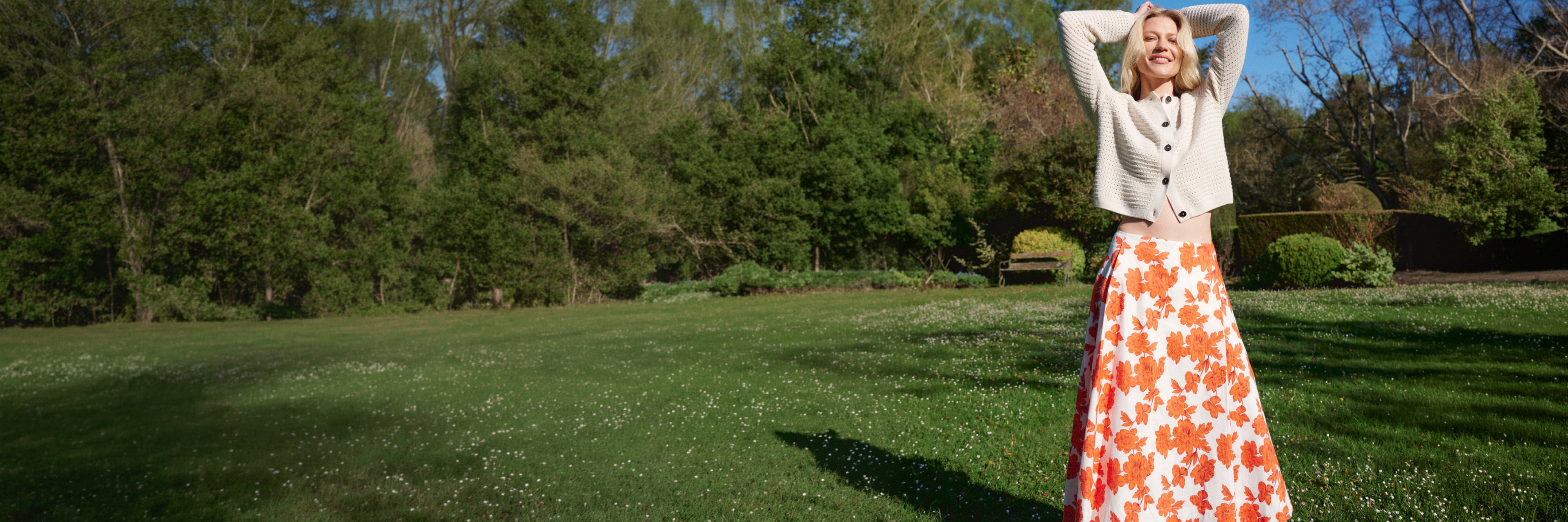 Woman in a floral skirt and white sweater standing in a grassy field with trees in the background