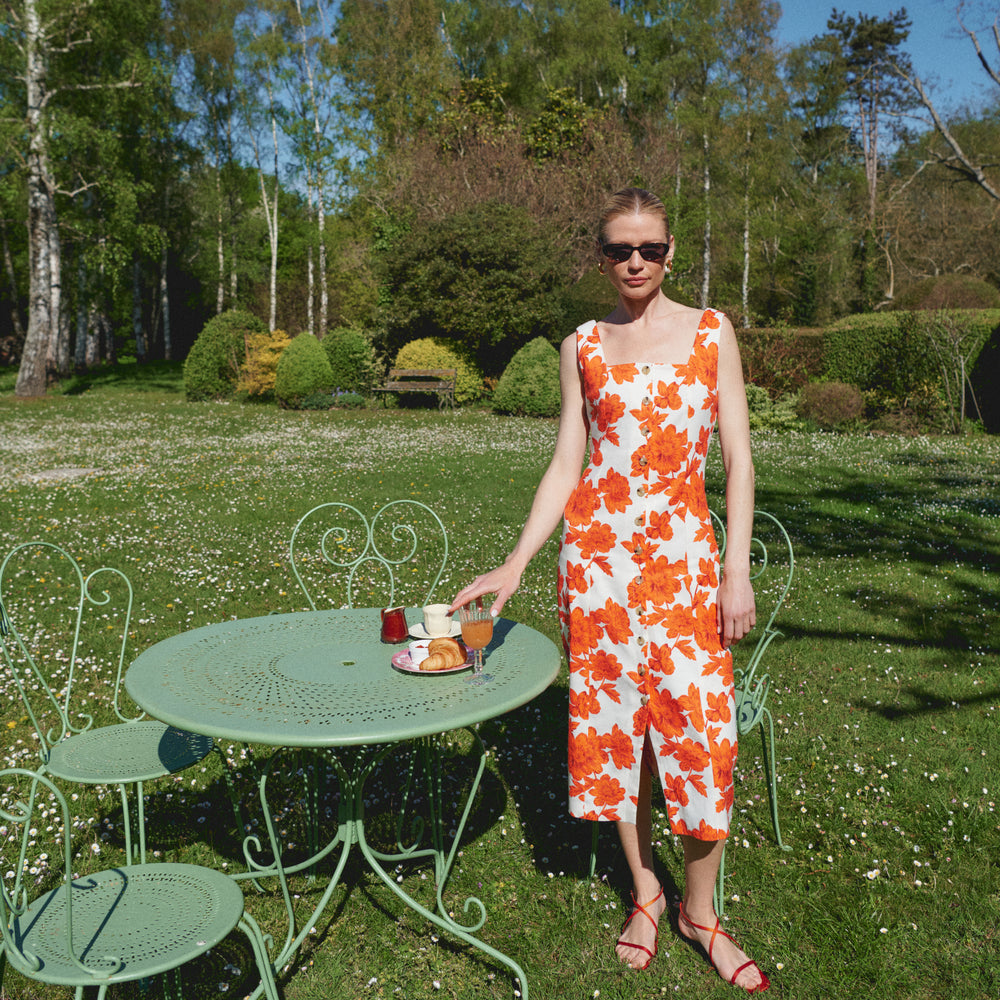 Woman in an orange and white floral dress standing next to a garden table with a tea set.