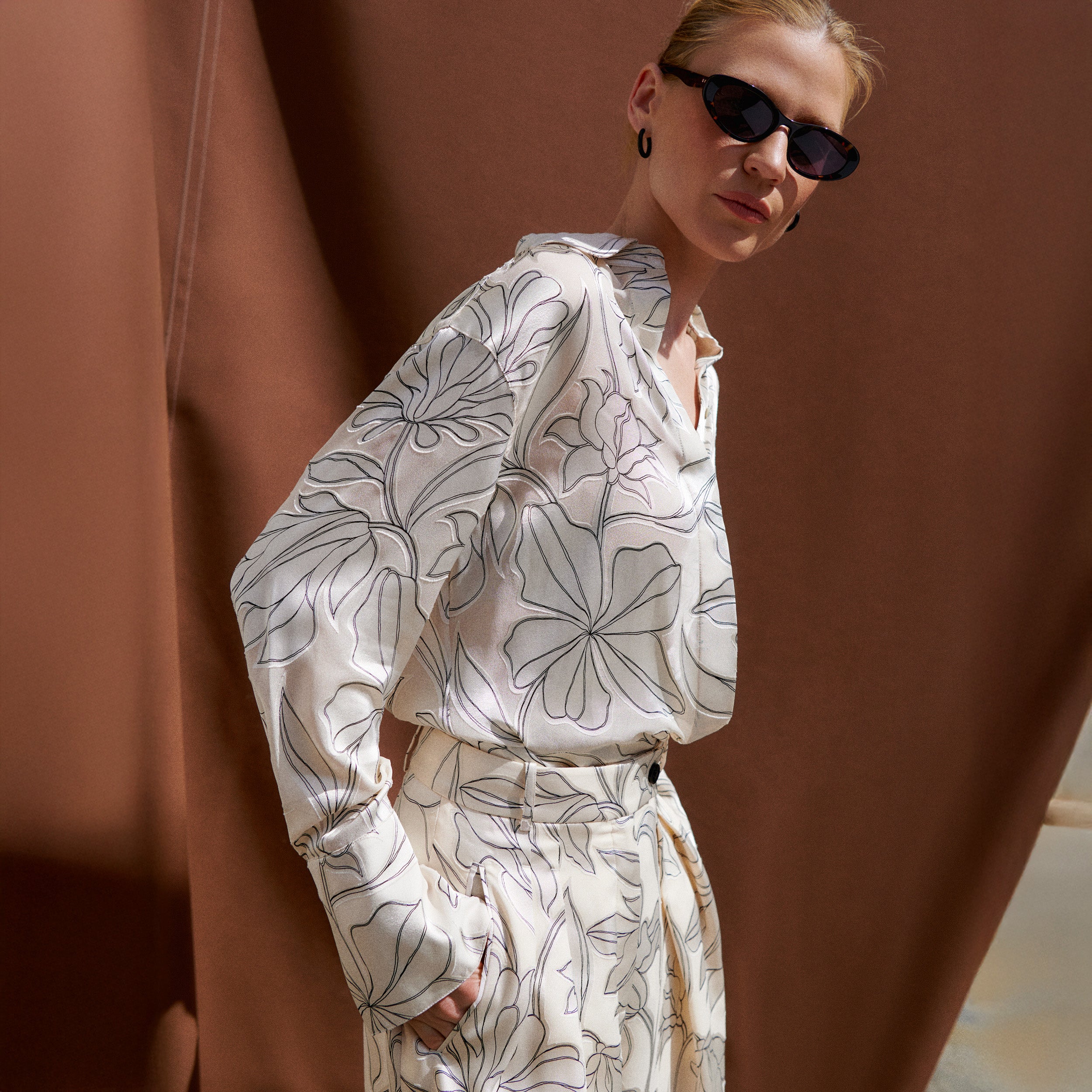 A woman wearing a cream-colored long-sleeve shirt and matching tie belt with a black floral line-art pattern, styled with black sunglasses, posing against a draped brown backdrop.