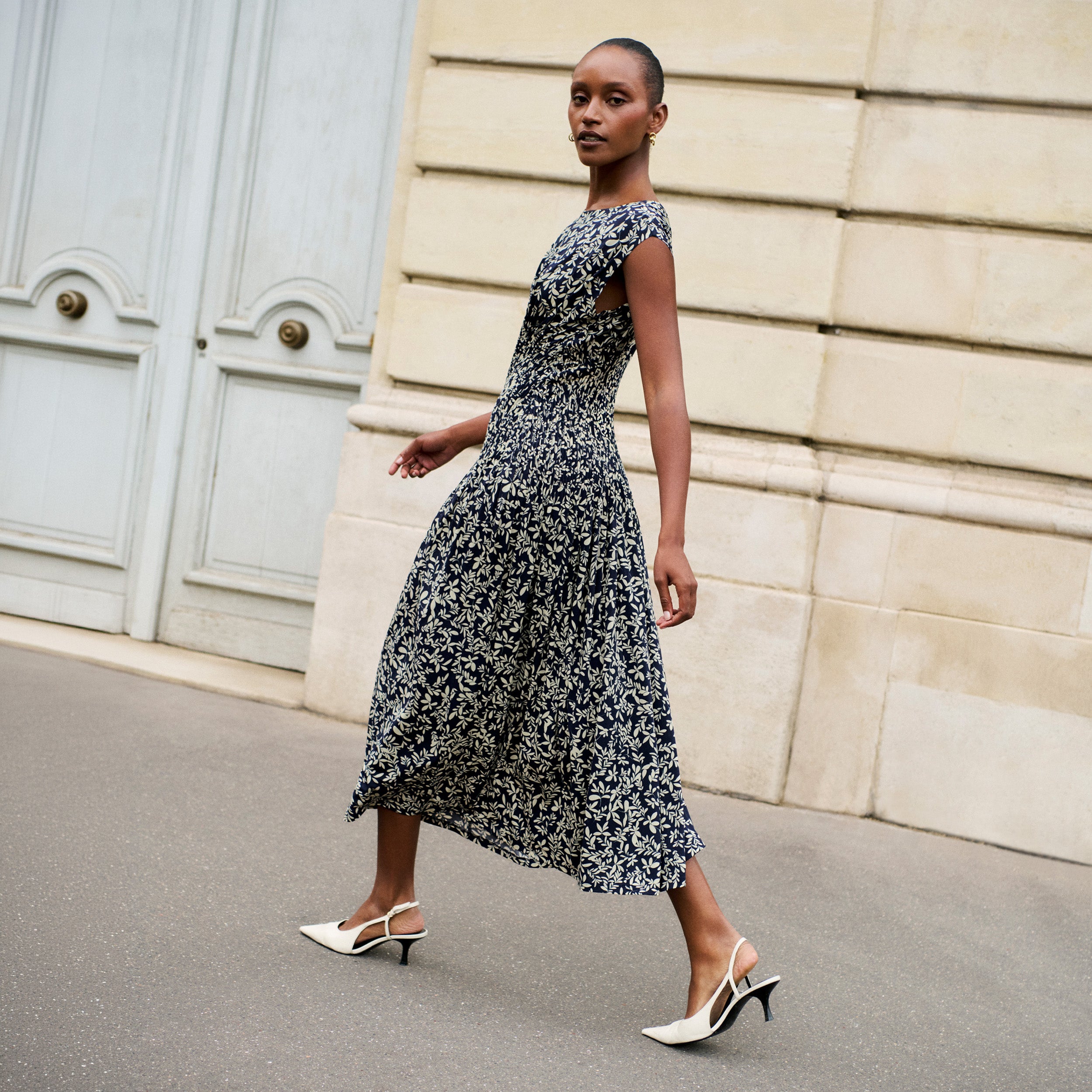 A woman walking outdoors in a sleeveless black midi dress with a white abstract floral pattern, paired with white slingback kitten heels, set against a beige stone wall and pale blue doors.