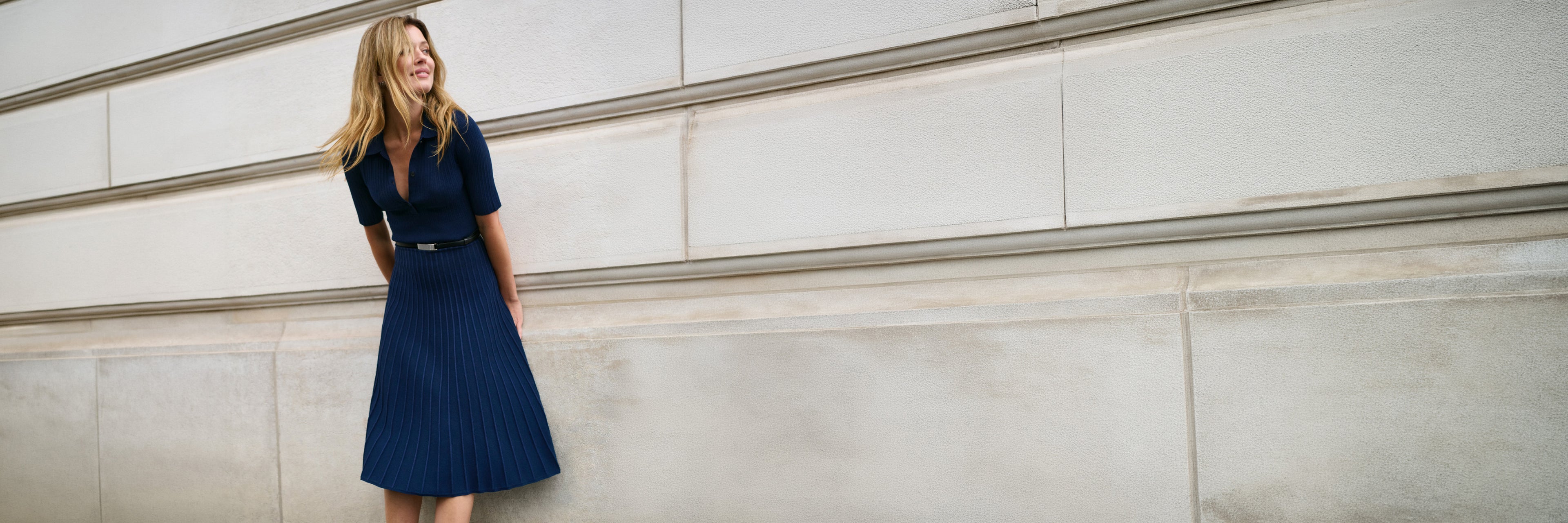 A woman wearing a dark navy blue pleated midi dress with a matching belt stands against a light gray stone wall.