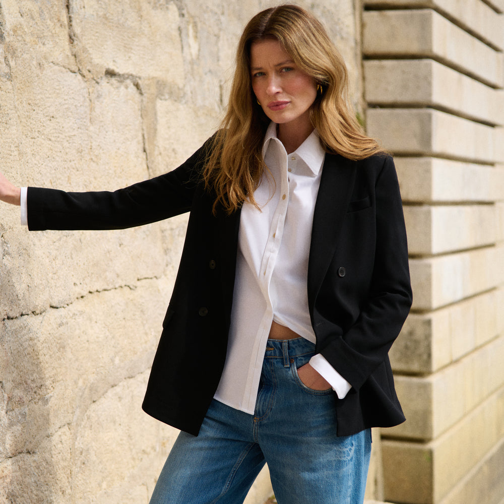 Woman wearing a black blazer, white shirt, and blue jeans standing against a stone wall.