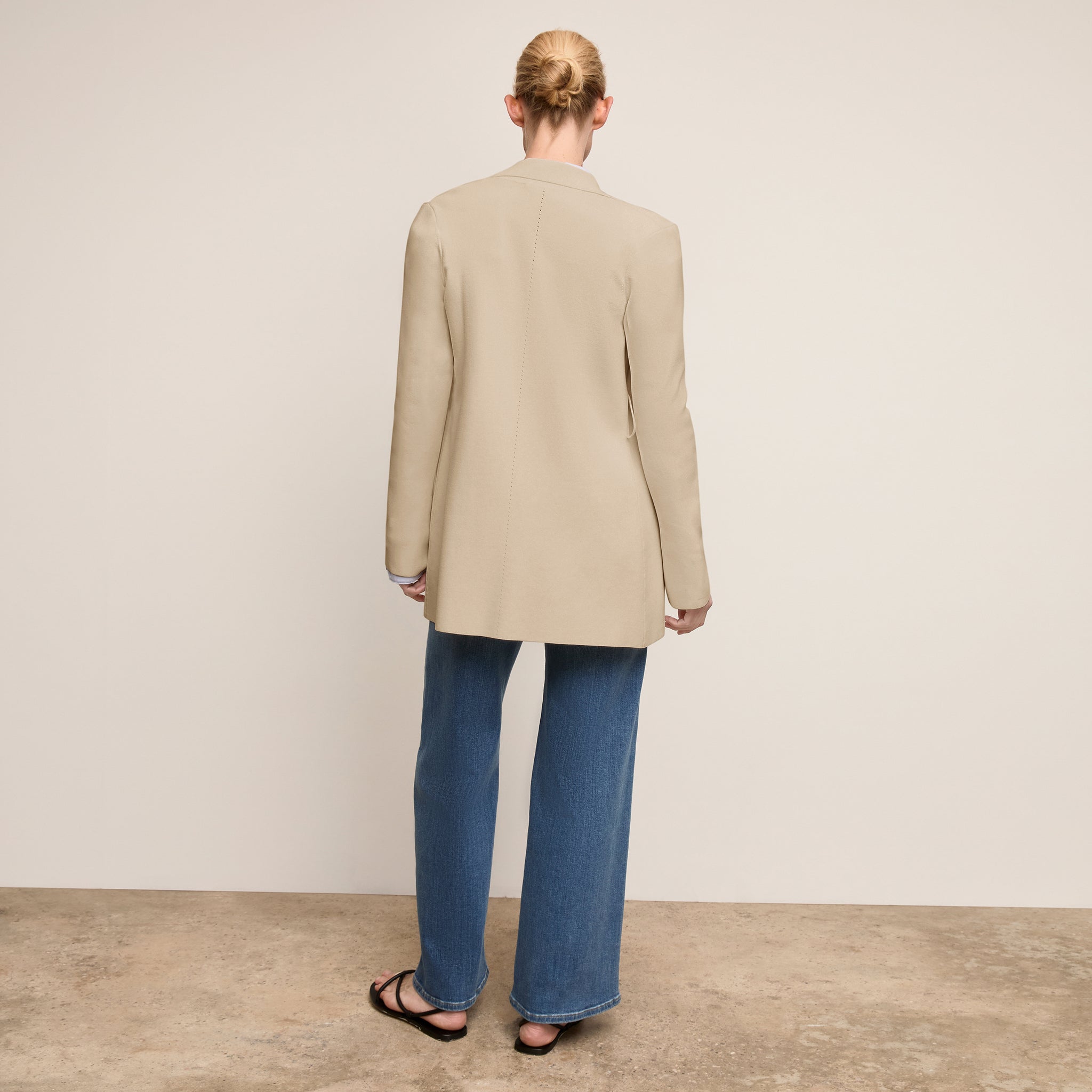 Person wearing a beige blazer and blue jeans standing against a plain background