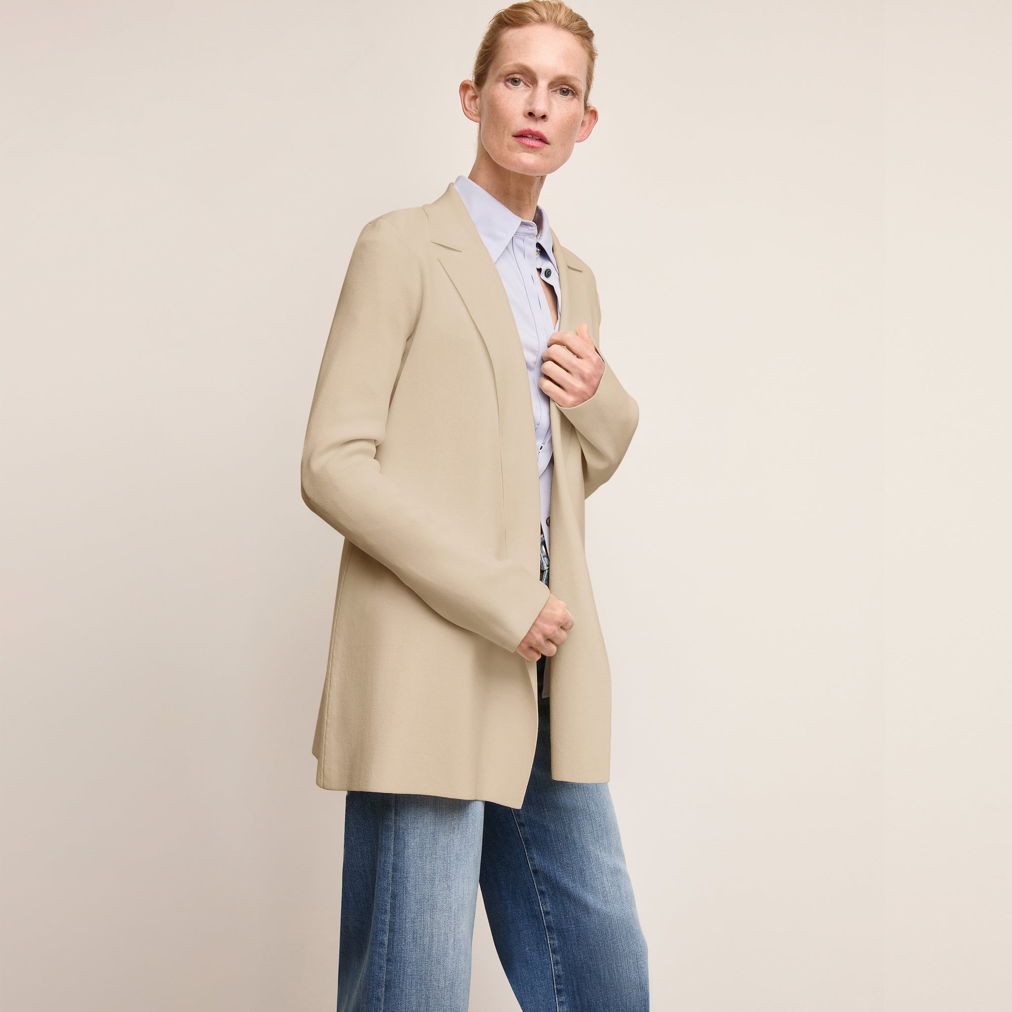 Woman wearing a beige blazer over a light blue shirt and jeans on a plain background 