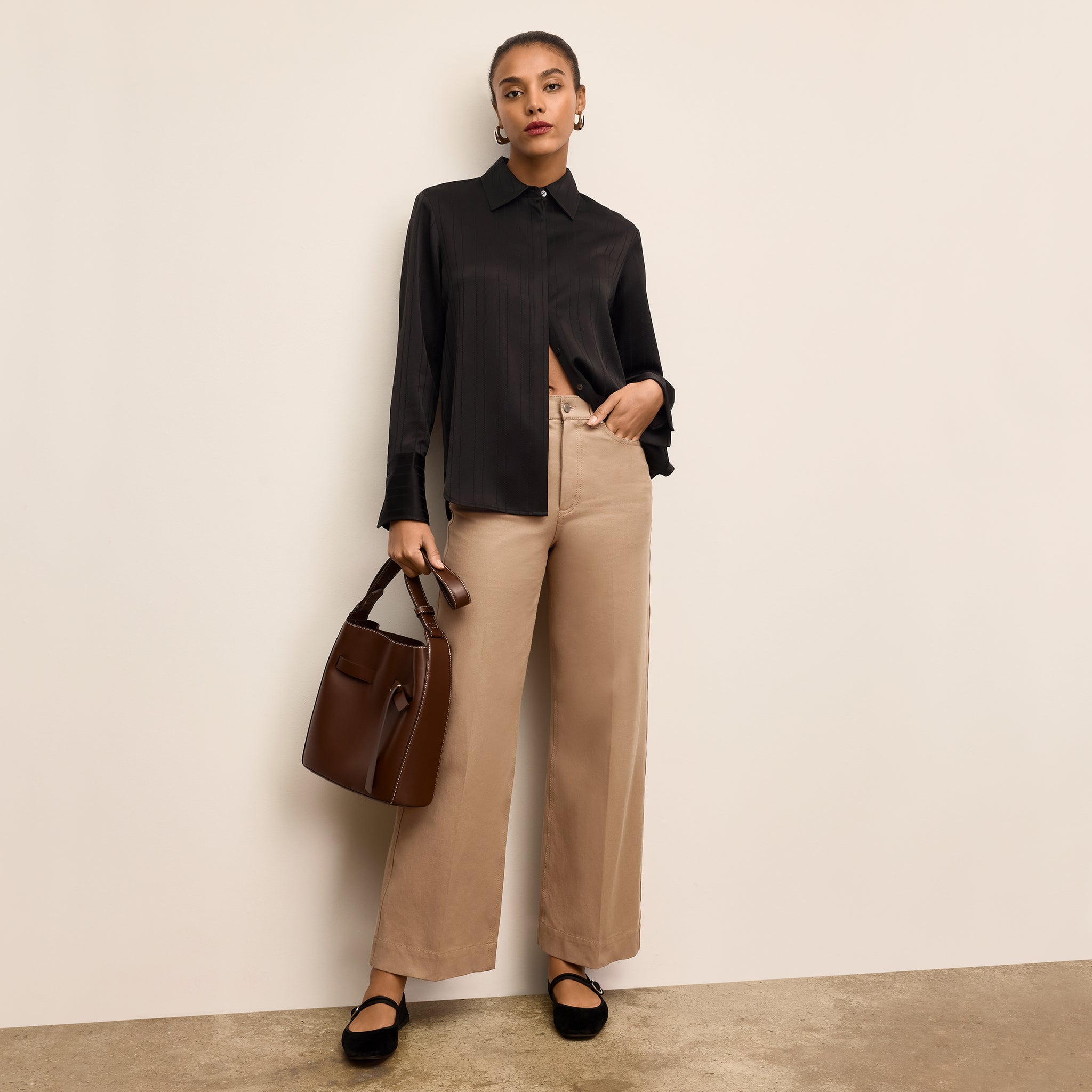 Woman wearing a black shirt and beige pants holding a brown handbag against a plain background