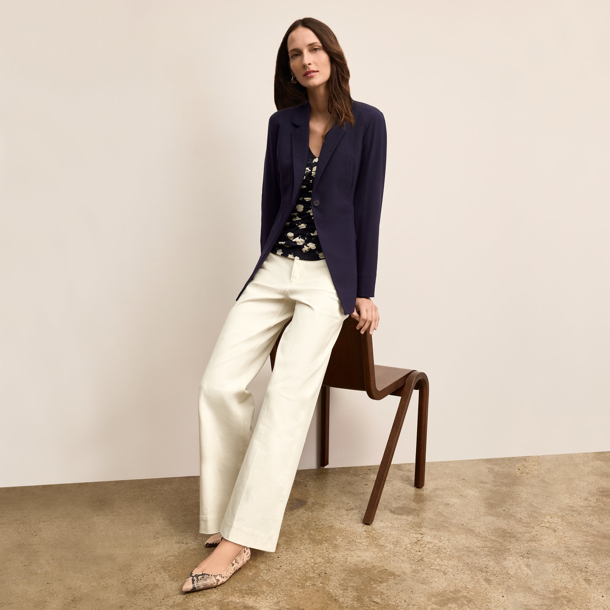 Woman sitting on a chair wearing a navy blazer, patterned top, and white pants against a plain background.