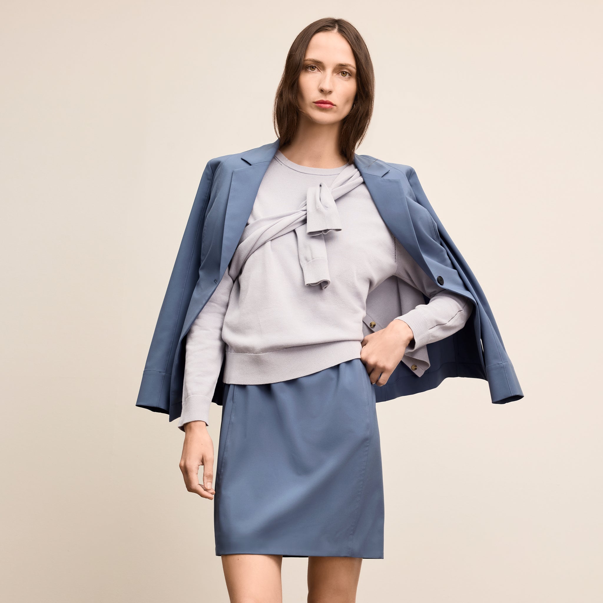 Woman wearing a light gray sweater with a bow, blue skirt, and blue blazer on a beige background