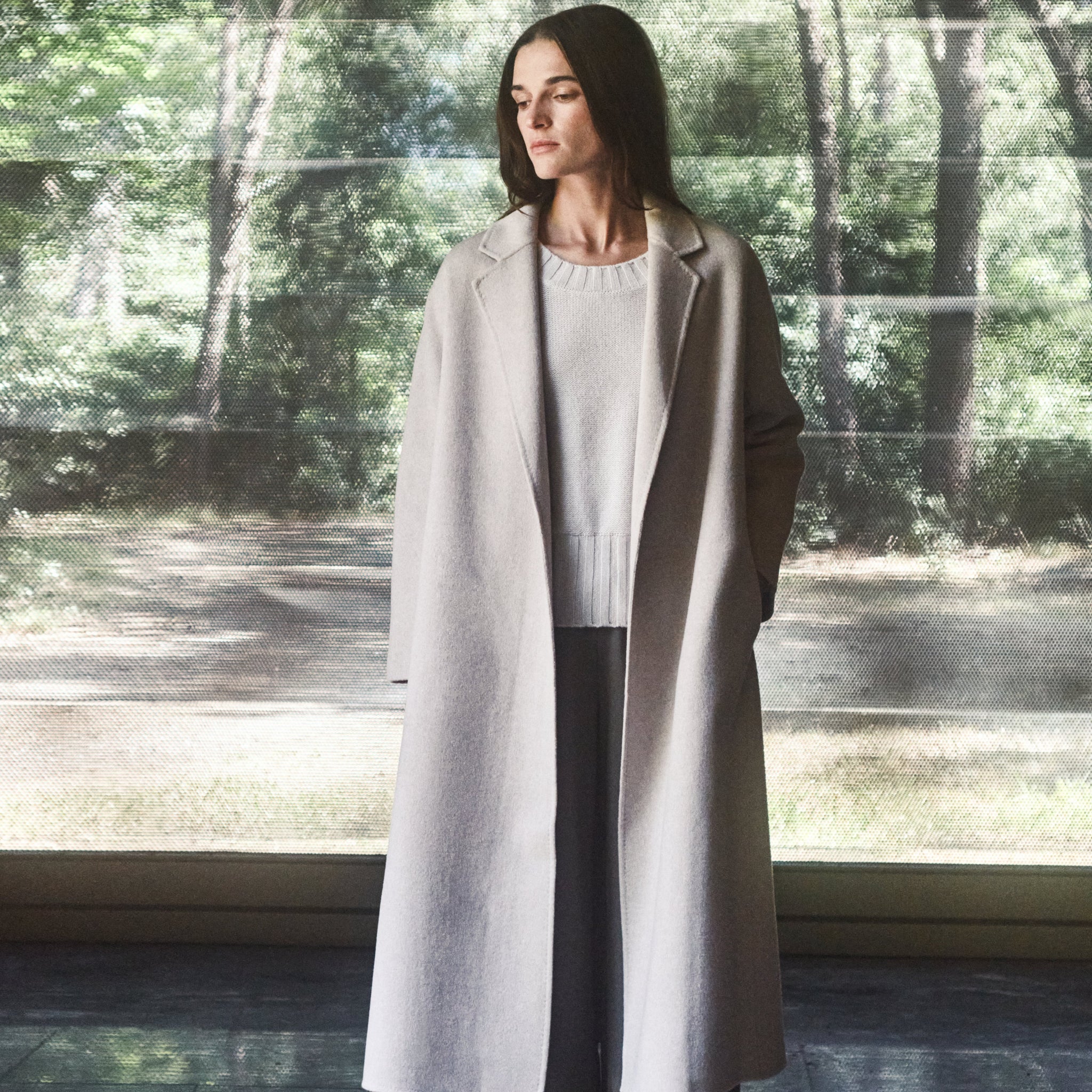Woman wearing a long coat standing in front of a reflective surface with a forest background