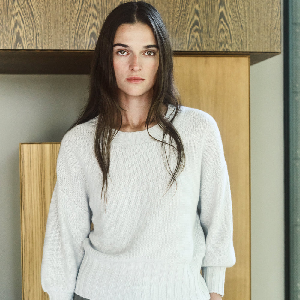 Woman wearing a white sweater and green pants standing indoors with wooden panels in the background