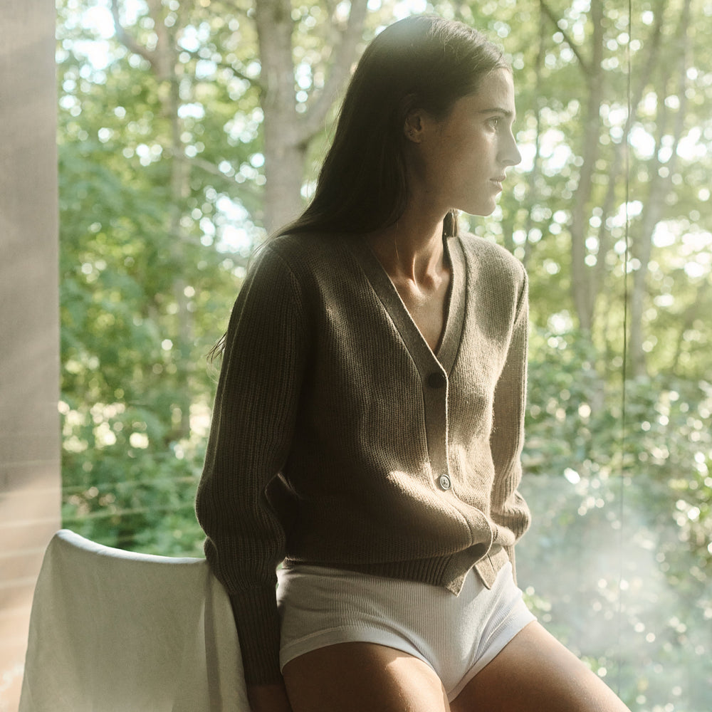 Woman in a brown sweater and white shorts standing in a natural setting with trees.