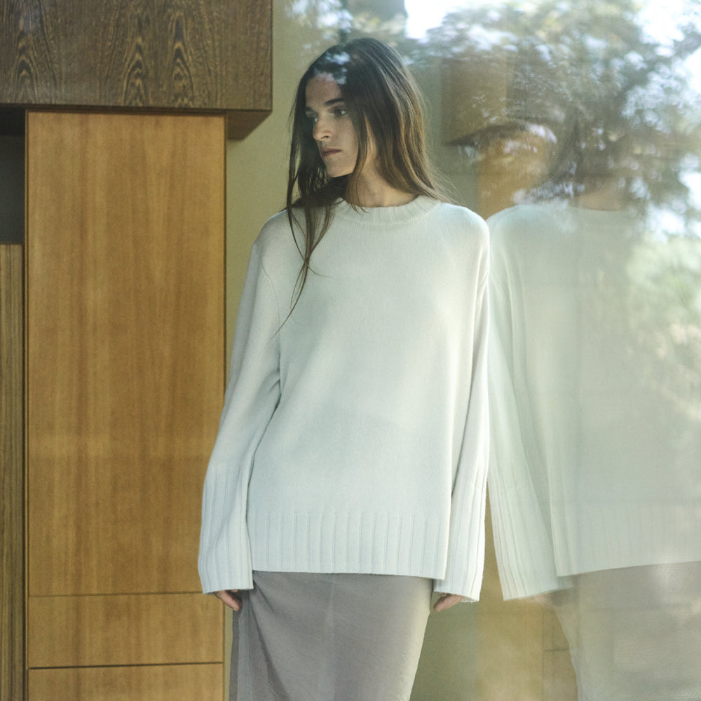 Woman wearing a light-colored sweater and pants standing in front of a glass door with wooden panels.