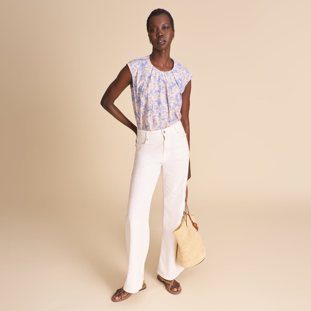 Woman wearing a floral top and white pants, holding a beige bag on a beige background