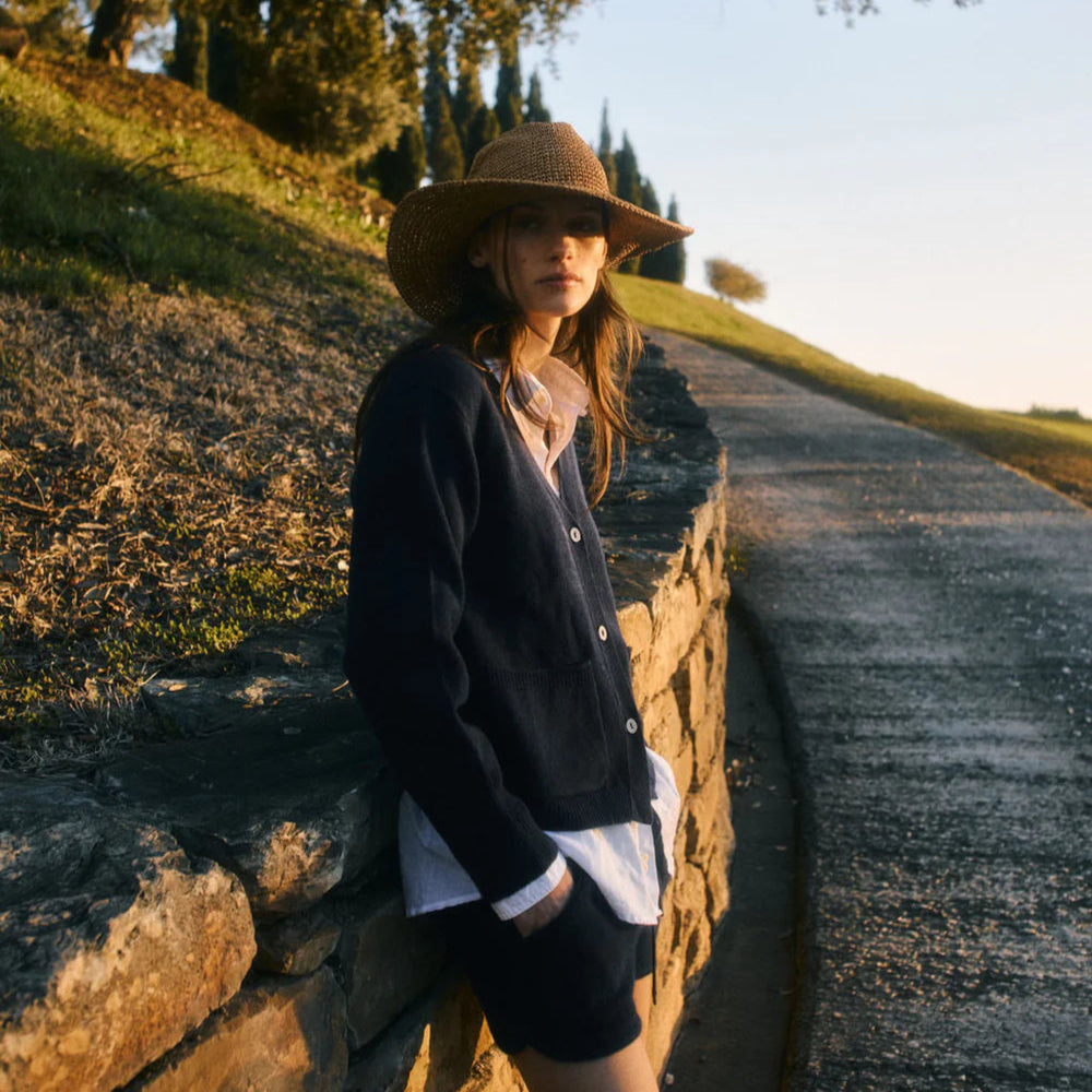 Woman wearing a wide-brimmed hat leaning against a stone wall by a road.