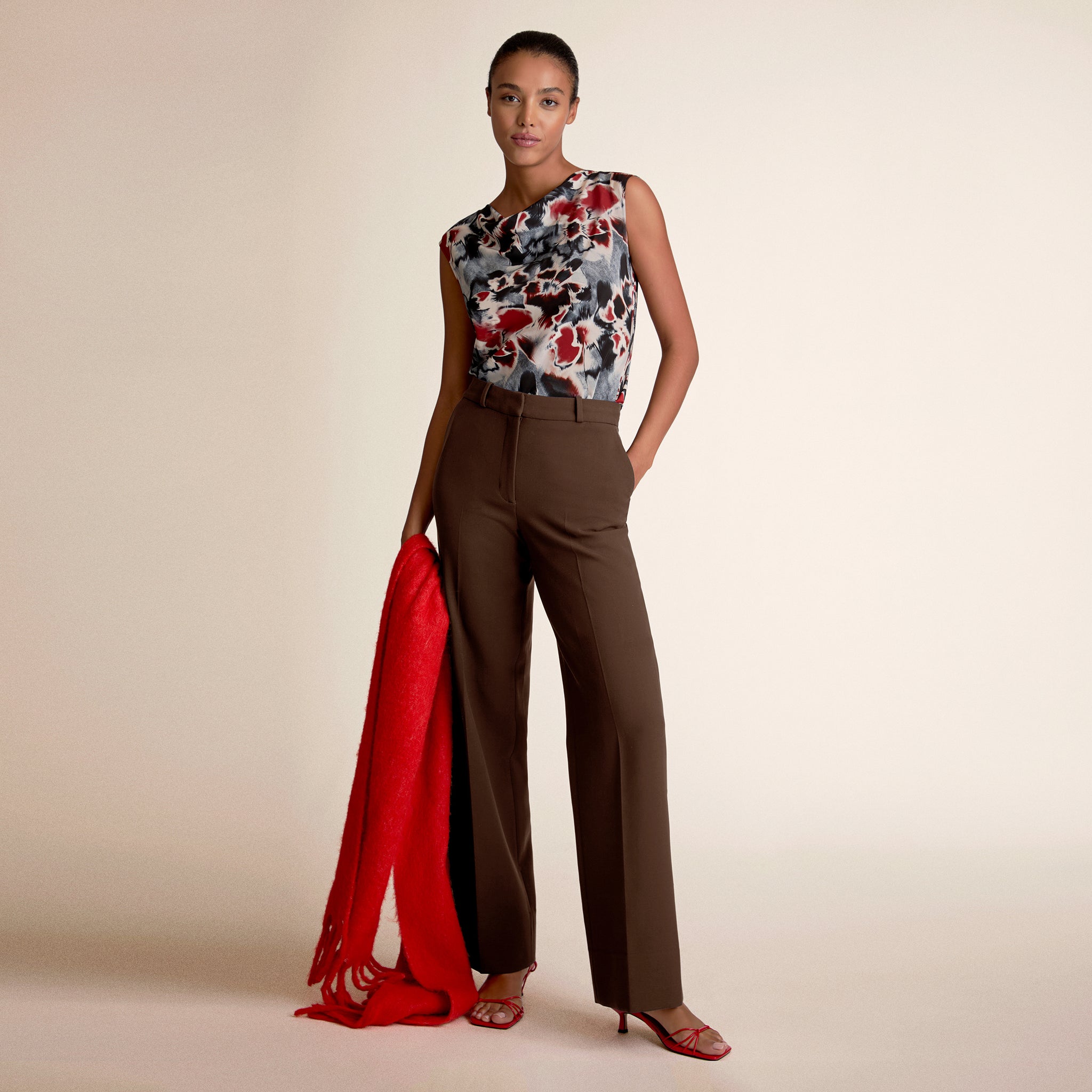 Woman wearing a floral top and brown pants with a red scarf, standing against a beige background 