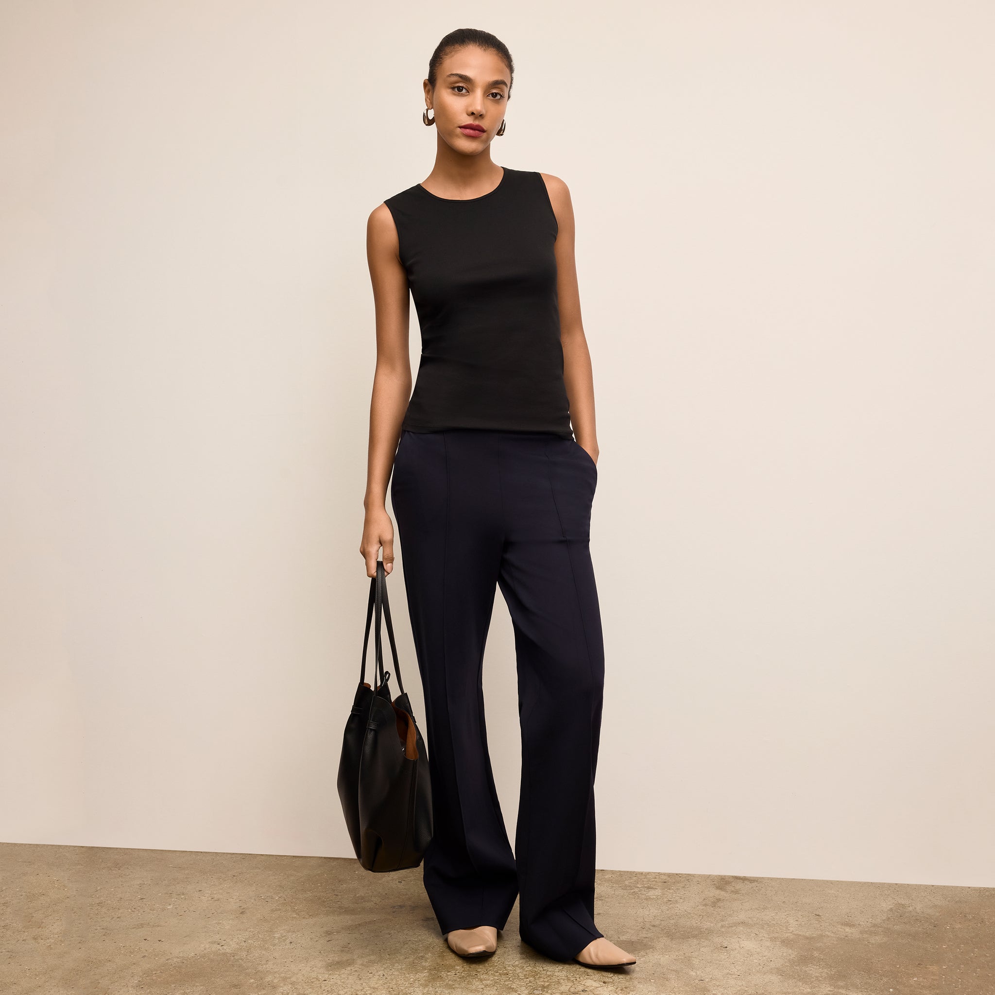 Woman wearing a black sleeveless top and pants holding a black handbag against a beige background