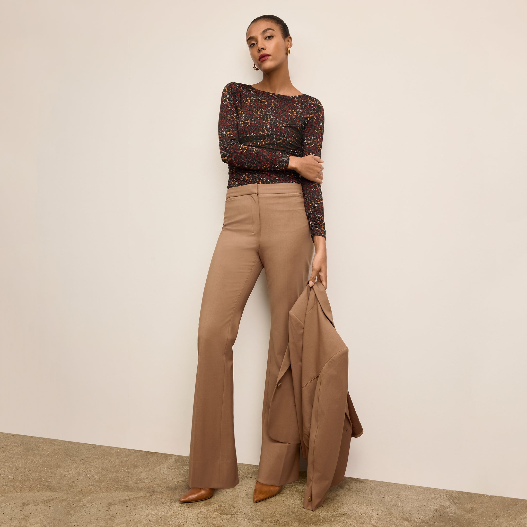 Woman wearing a patterned top and beige pants holding a matching jacket against a neutral background