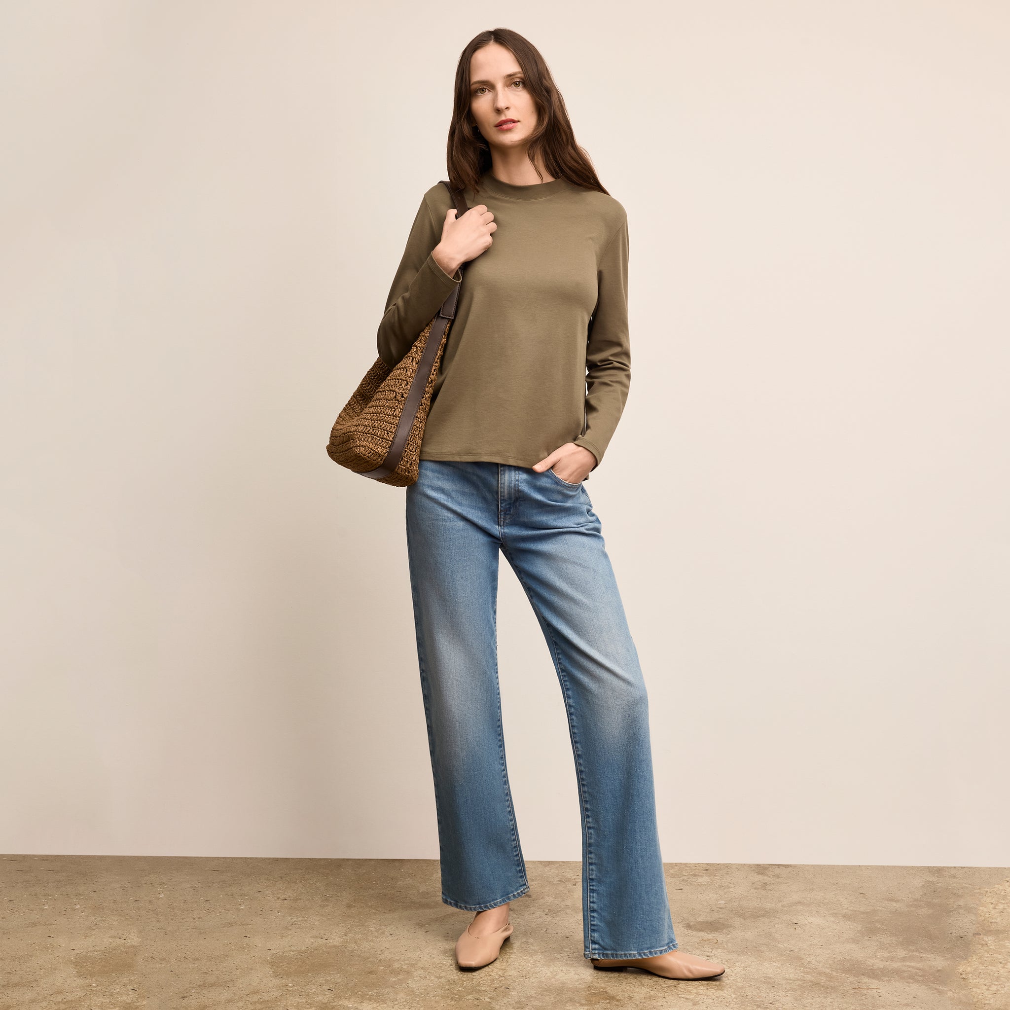 Woman wearing a green sweater and blue jeans holding a woven bag against a beige background 