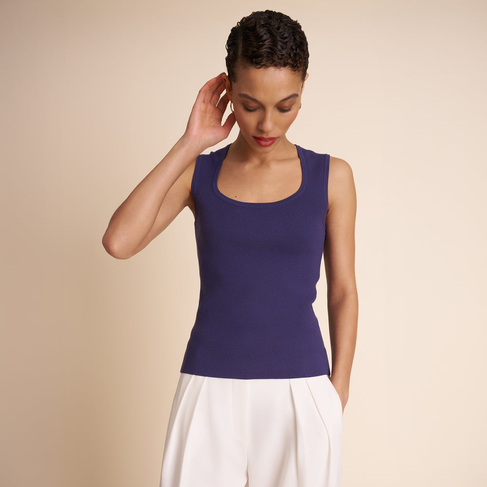 Woman wearing a navy blue tank top and white pants against a beige background