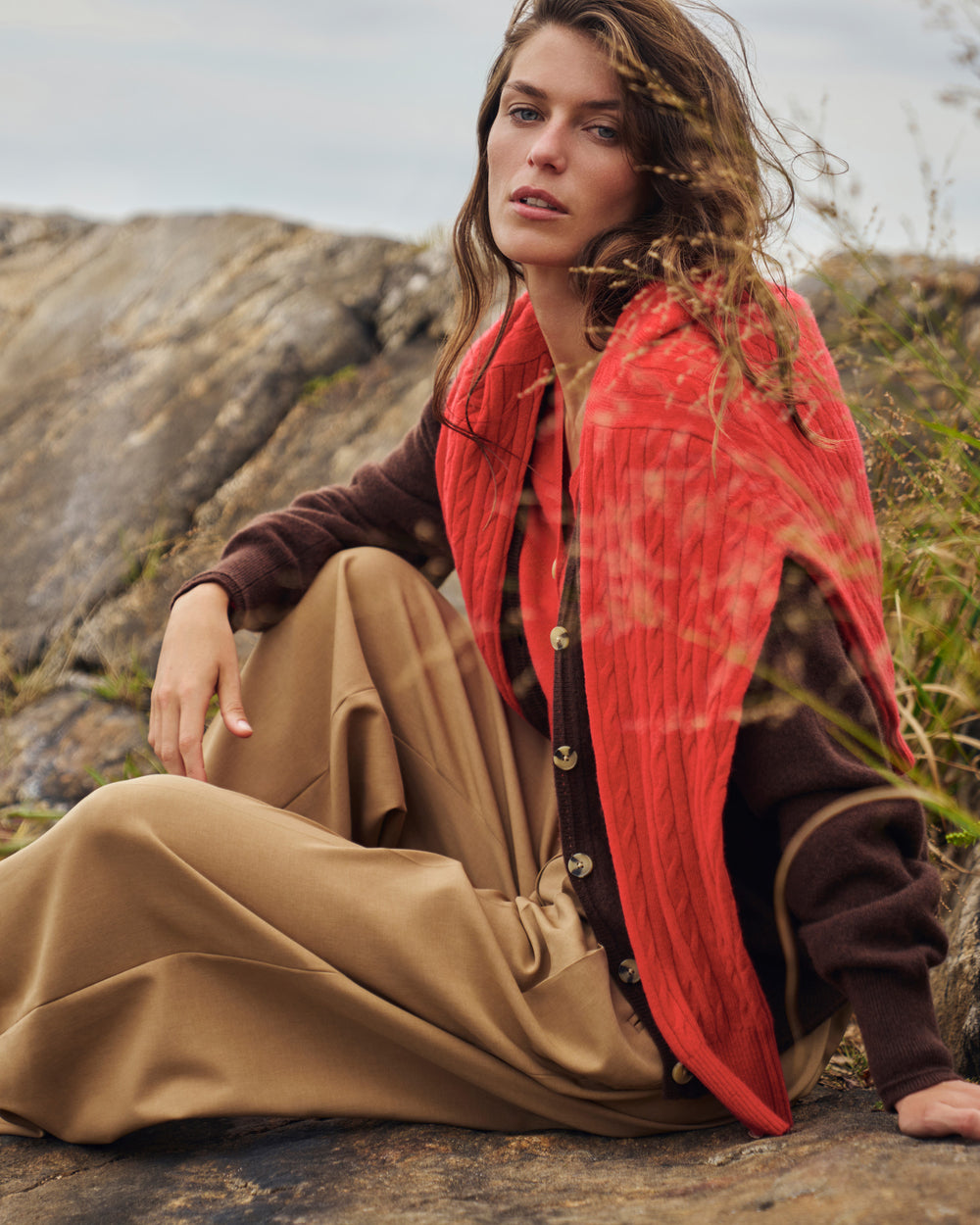 A woman sits outdoors on a rocky surface wearing a rust-red textured wrap/shawl over a brown cardigan and tan wide-leg pants.
