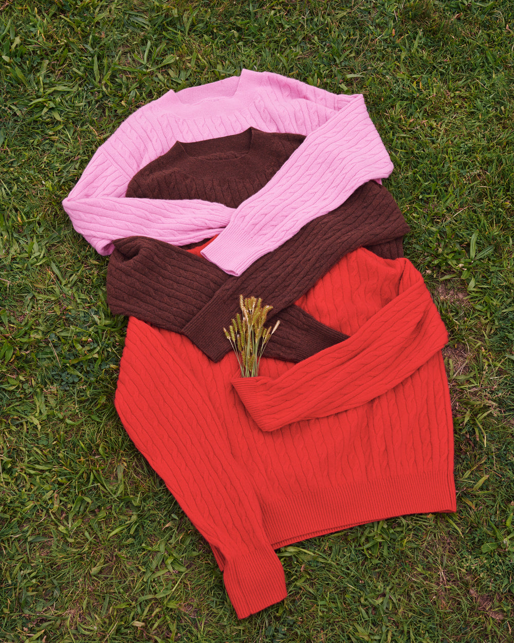 Three ribbed knit sweaters in pink, brown, and red are arranged in a stack on green grass, with a small sprig of dried grass placed on top.