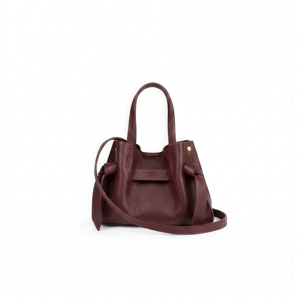 A small pebbled leather handbag in deep burgundy, featuring dual top handles, a detachable shoulder strap, and a front leather detail with a small gold rivet, set against a plain white background.