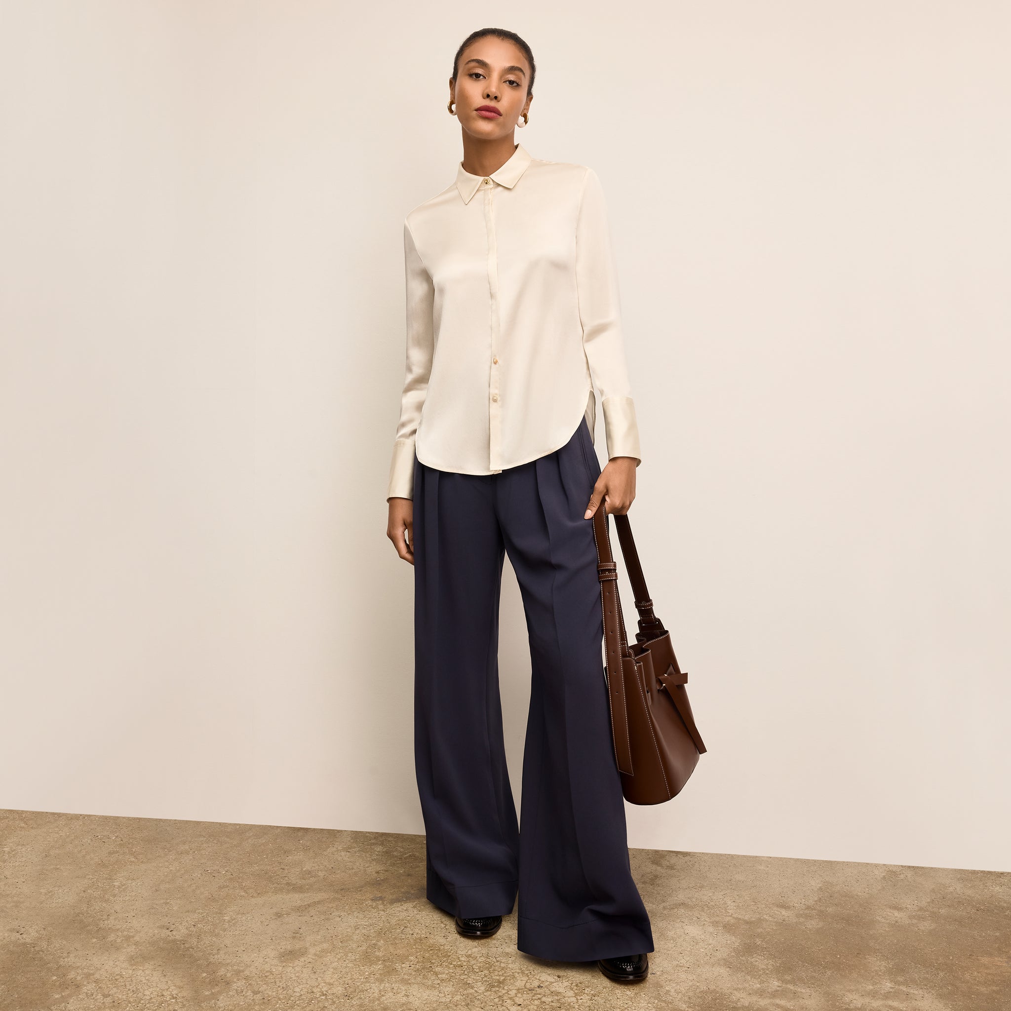 Woman wearing a white blouse and navy pants, holding a brown handbag against a plain background 