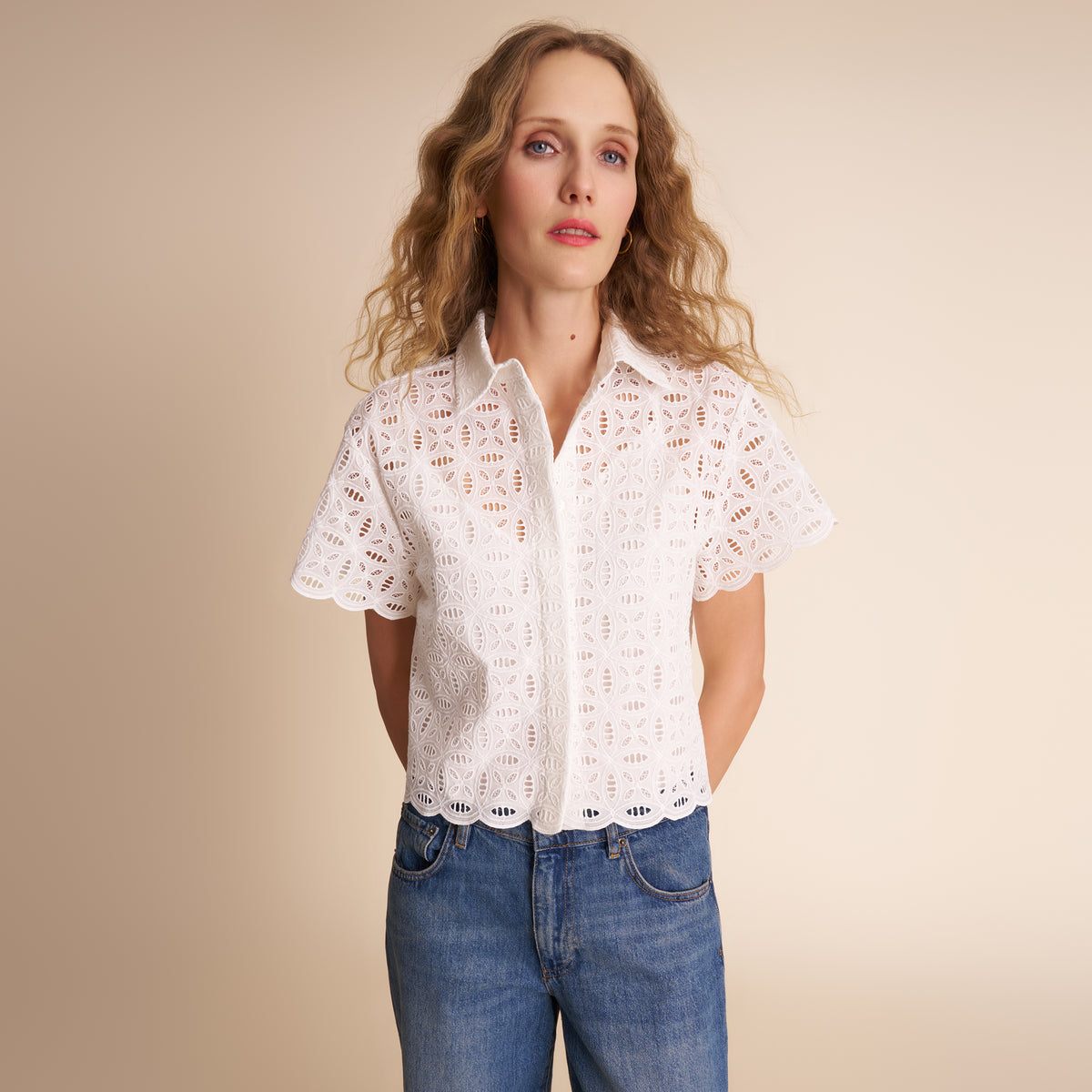 Woman wearing a white lace blouse and blue jeans against a beige background