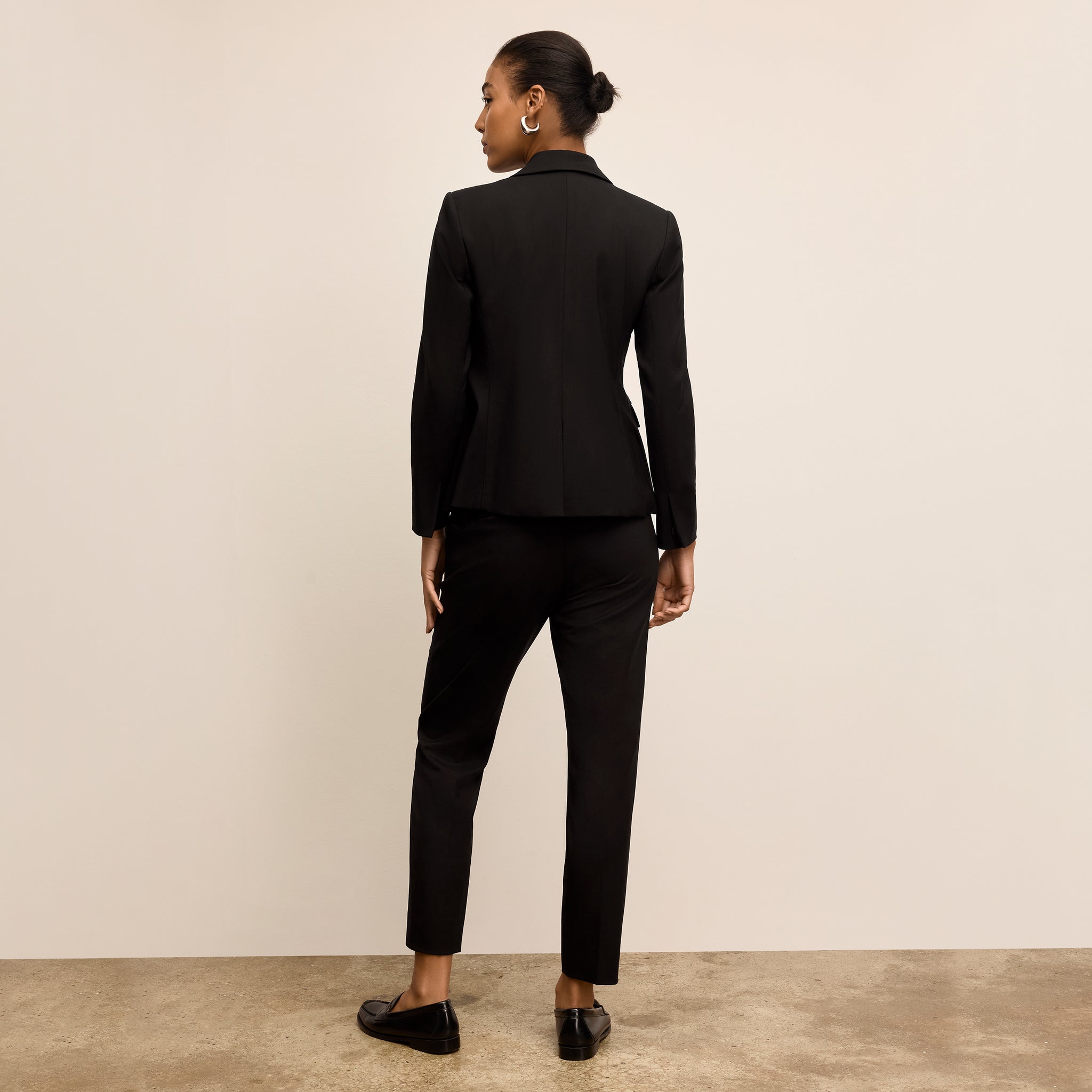 Person wearing a black suit standing against a beige wall.