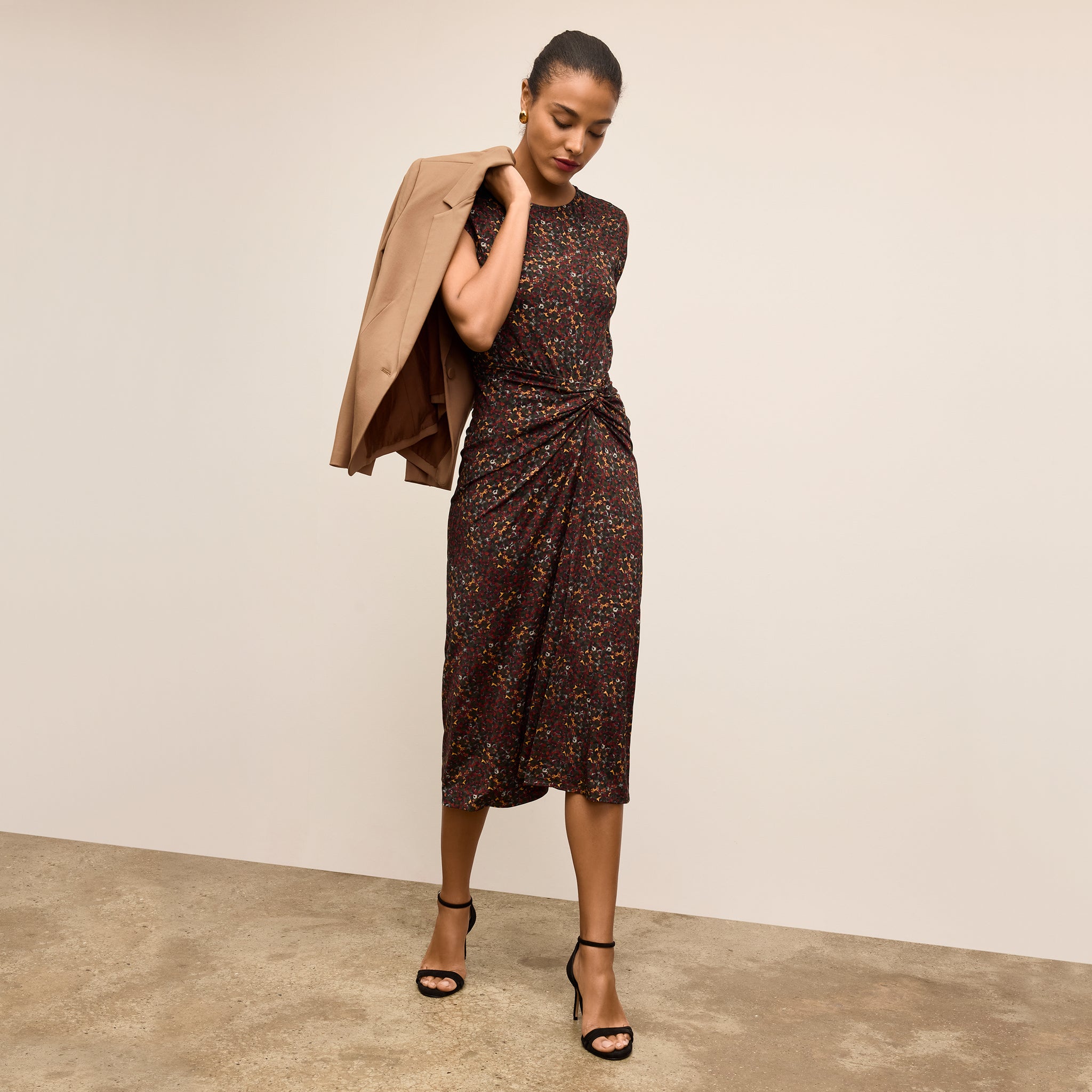 Woman wearing a patterned dress with a beige coat over her shoulder against a plain background