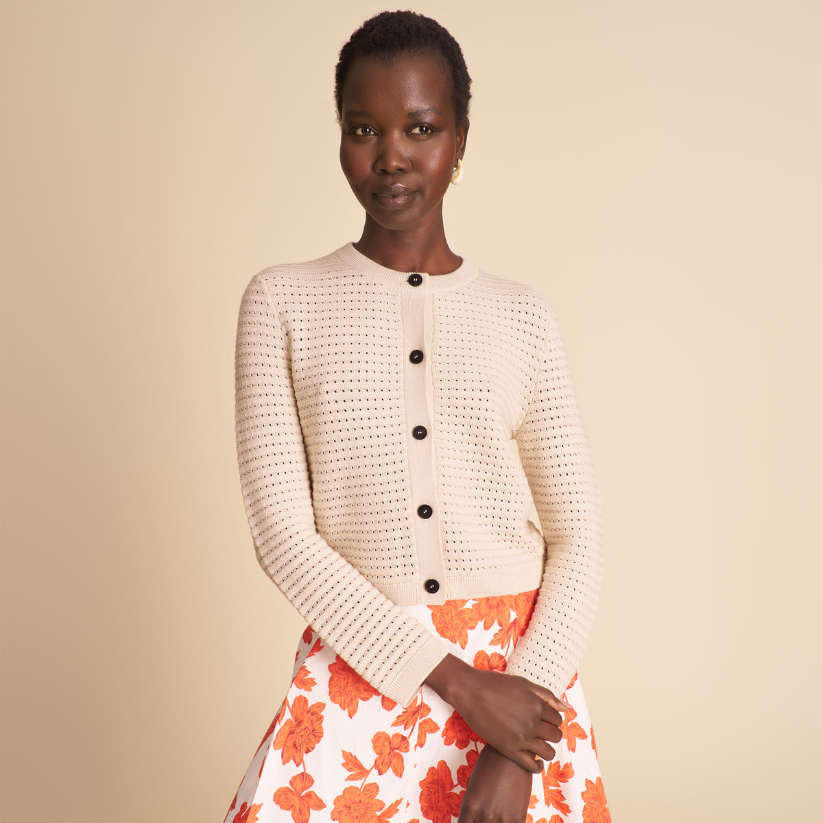Woman wearing a beige knitted cardigan with black buttons and an orange floral skirt on a beige background