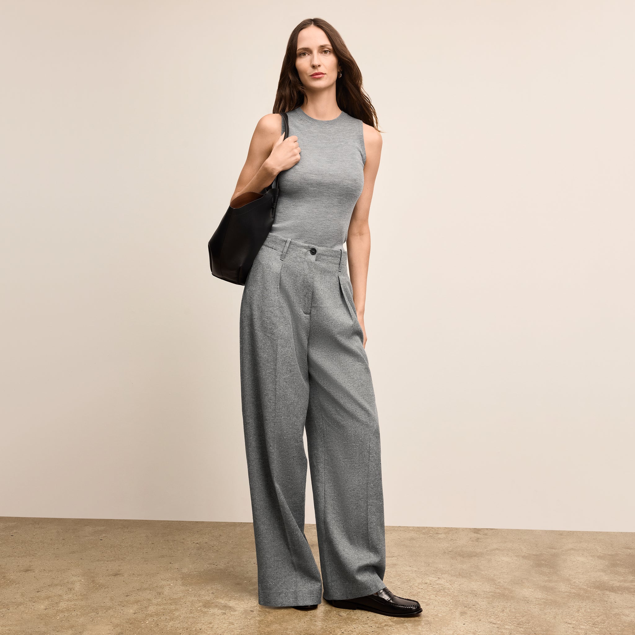 Woman wearing a gray sleeveless top and wide-leg pants holding a black handbag against a beige background