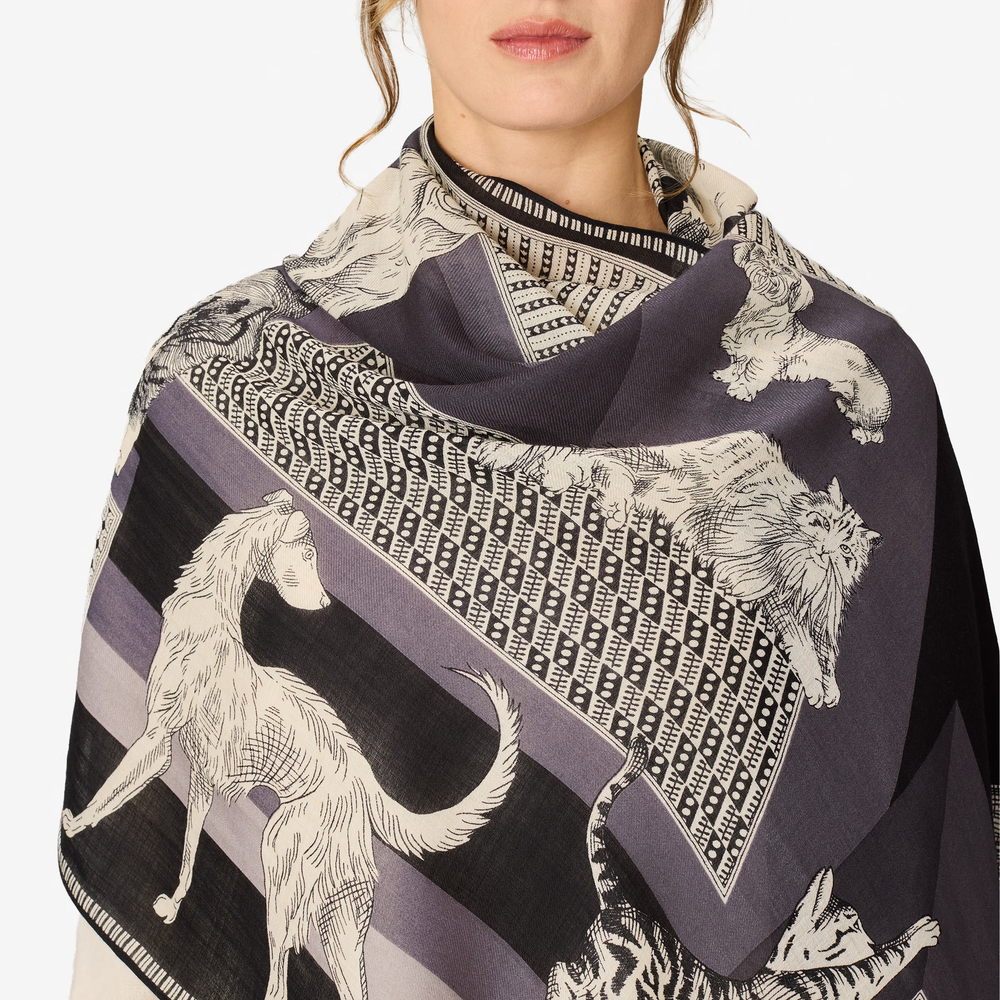 A close-up of a model wearing a dark patterned shawl with illustrations of dogs, cats, and geometric motifs in black, gray, and off-white.
