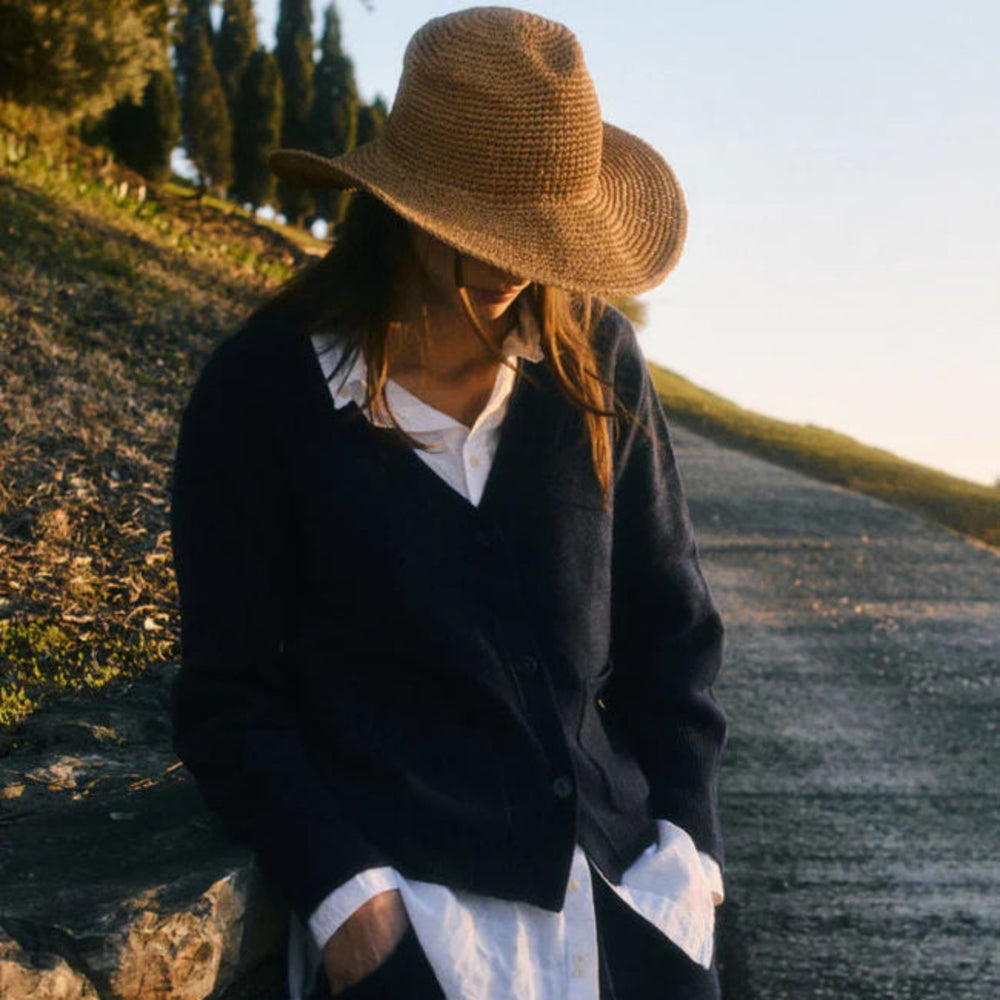 Person wearing a straw hat and dark jacket standing by a stone wall with a scenic background.