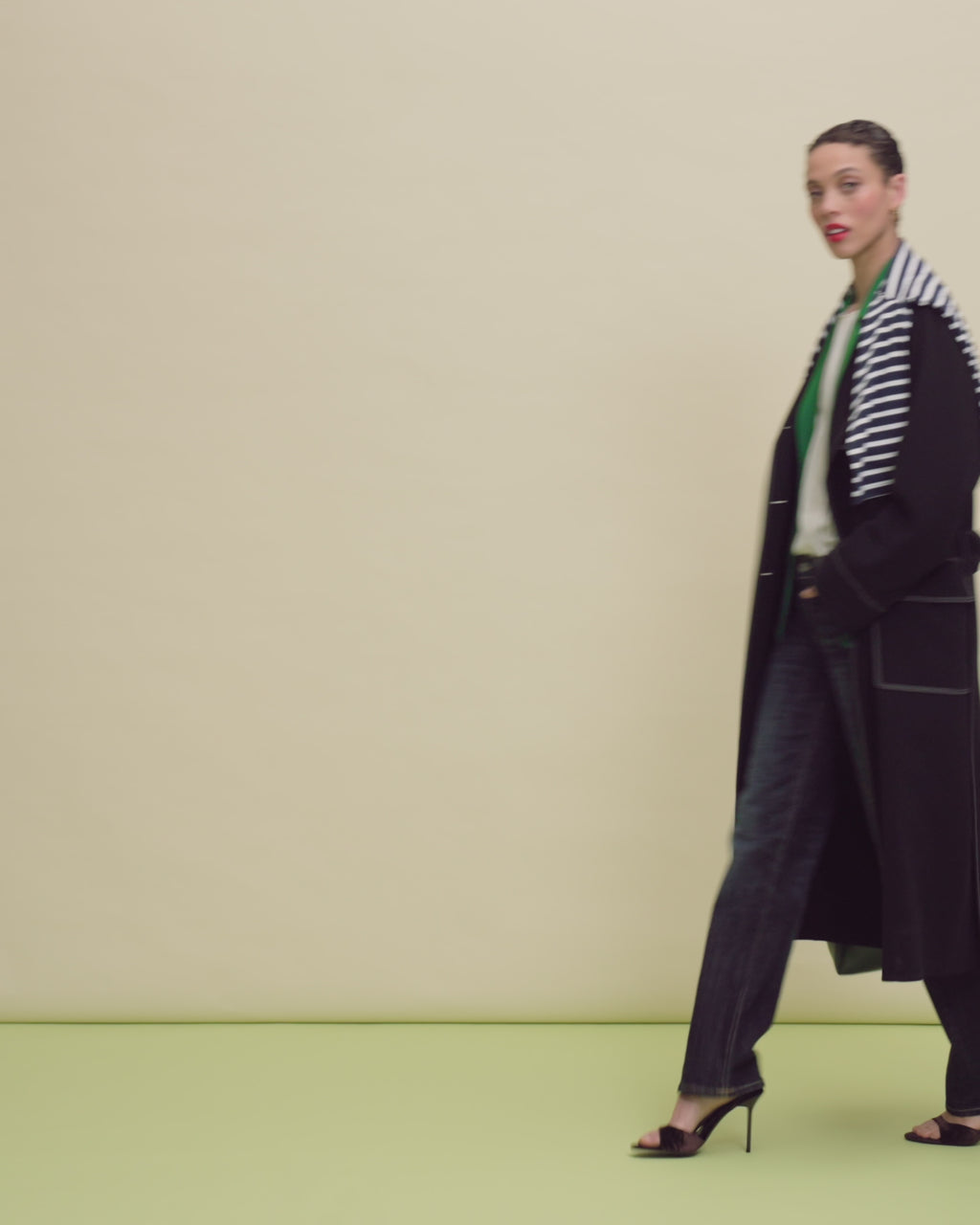 Woman wearing a green blazer, striped shirt, and blue jeans on a beige background.