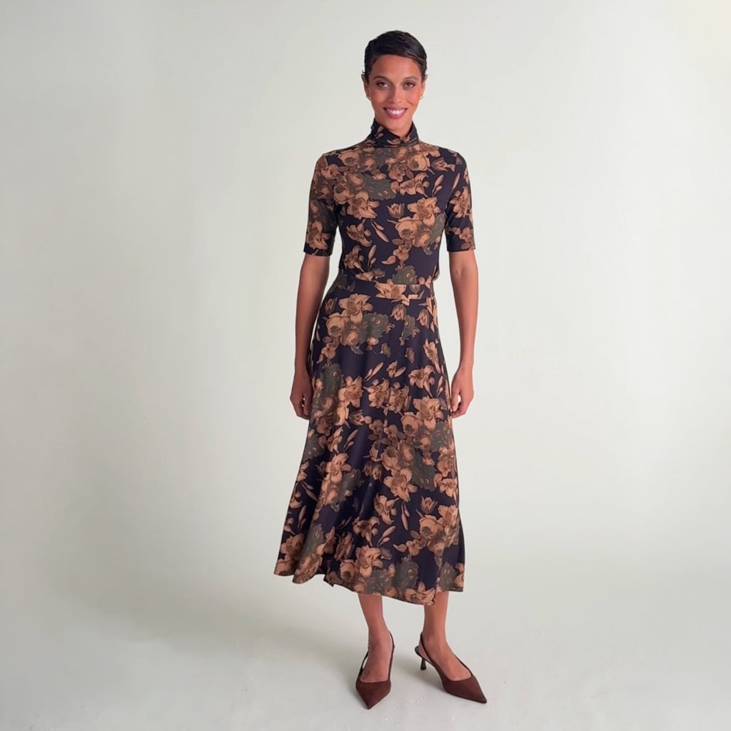 Video of a woman modeling a short-sleeved mock-neck midi dress with a dark navy blue base and a warm brown floral pattern, paired with dark brown pointed-toe slingback heels,