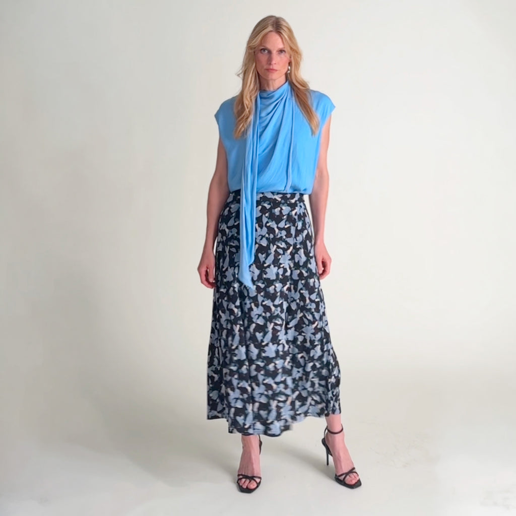 Woman standing wearing a light blue top with a scarf drape neckline and a navy and light blue printed maxi skirt