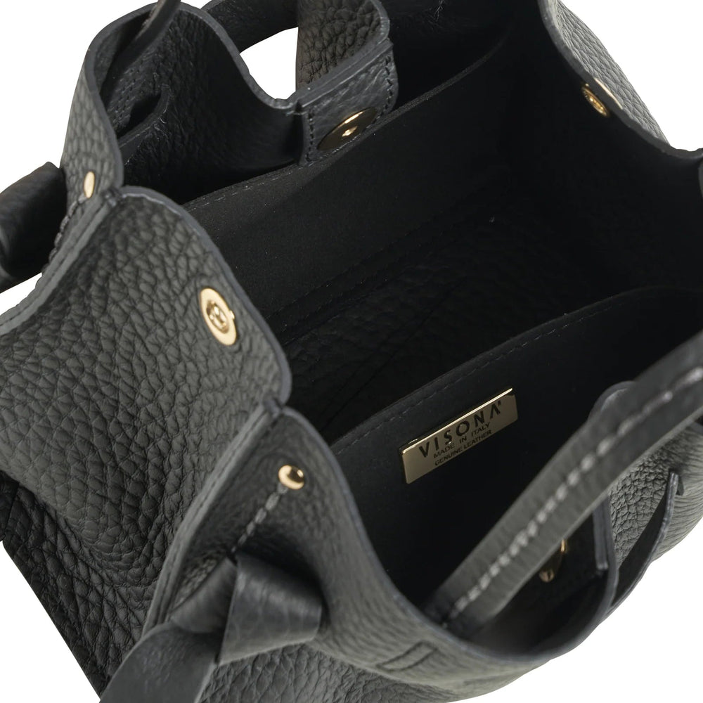 Close-up of a black pebbled leather handbag interior with a visible gold-tone metal plaque reading 'visona' on the inside wall, showing the black lining and part of the dark exterior strap against a white background.